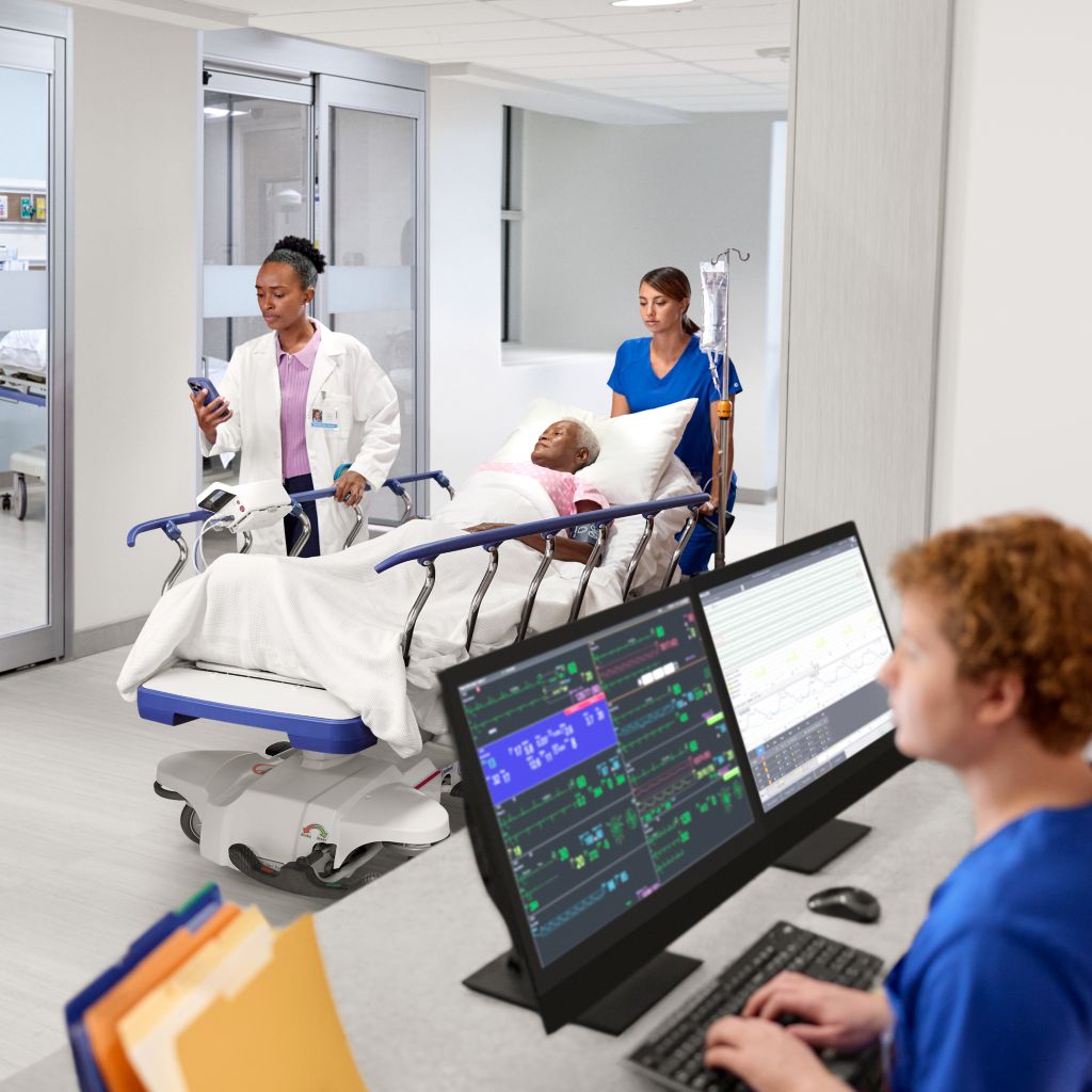 Clinician at central station while other clinicians wheel a patient down the hallway