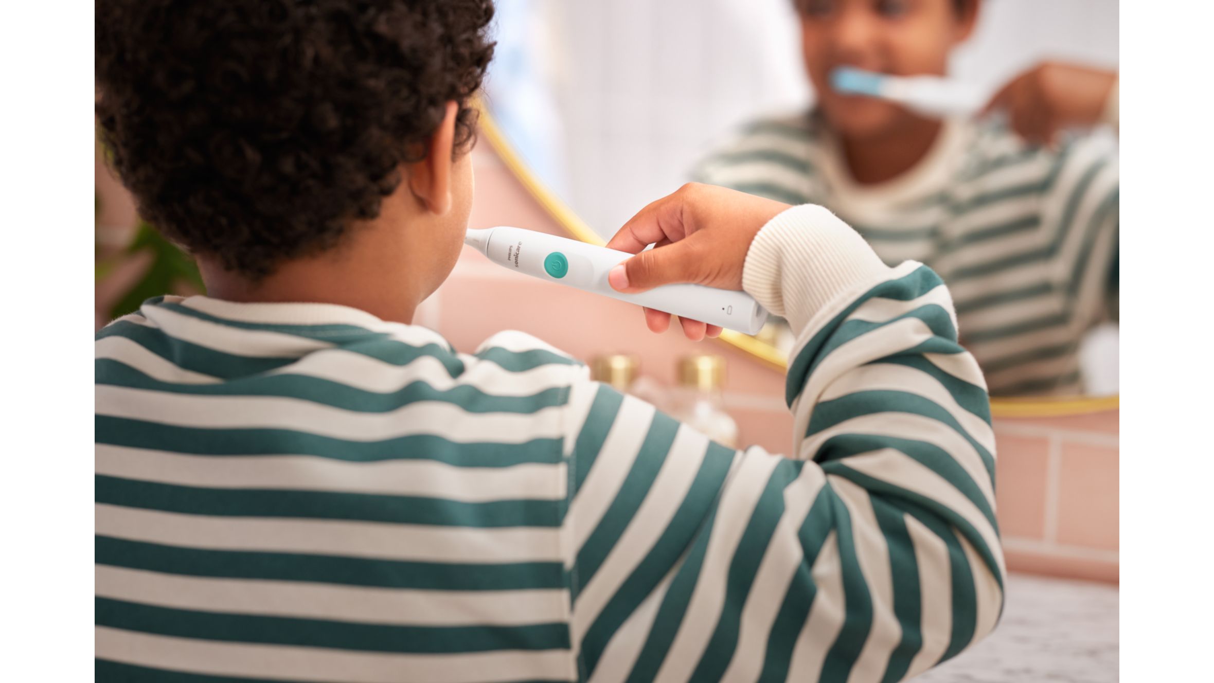 A child in a green and white striped top is shown from behind while brushing their teeth in the mirror.