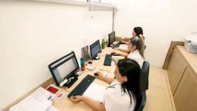 Equipe administrativa de saúde trabalhando no sistema Tasy utilizando computadores desktop em um espaço de escritório compartilhado