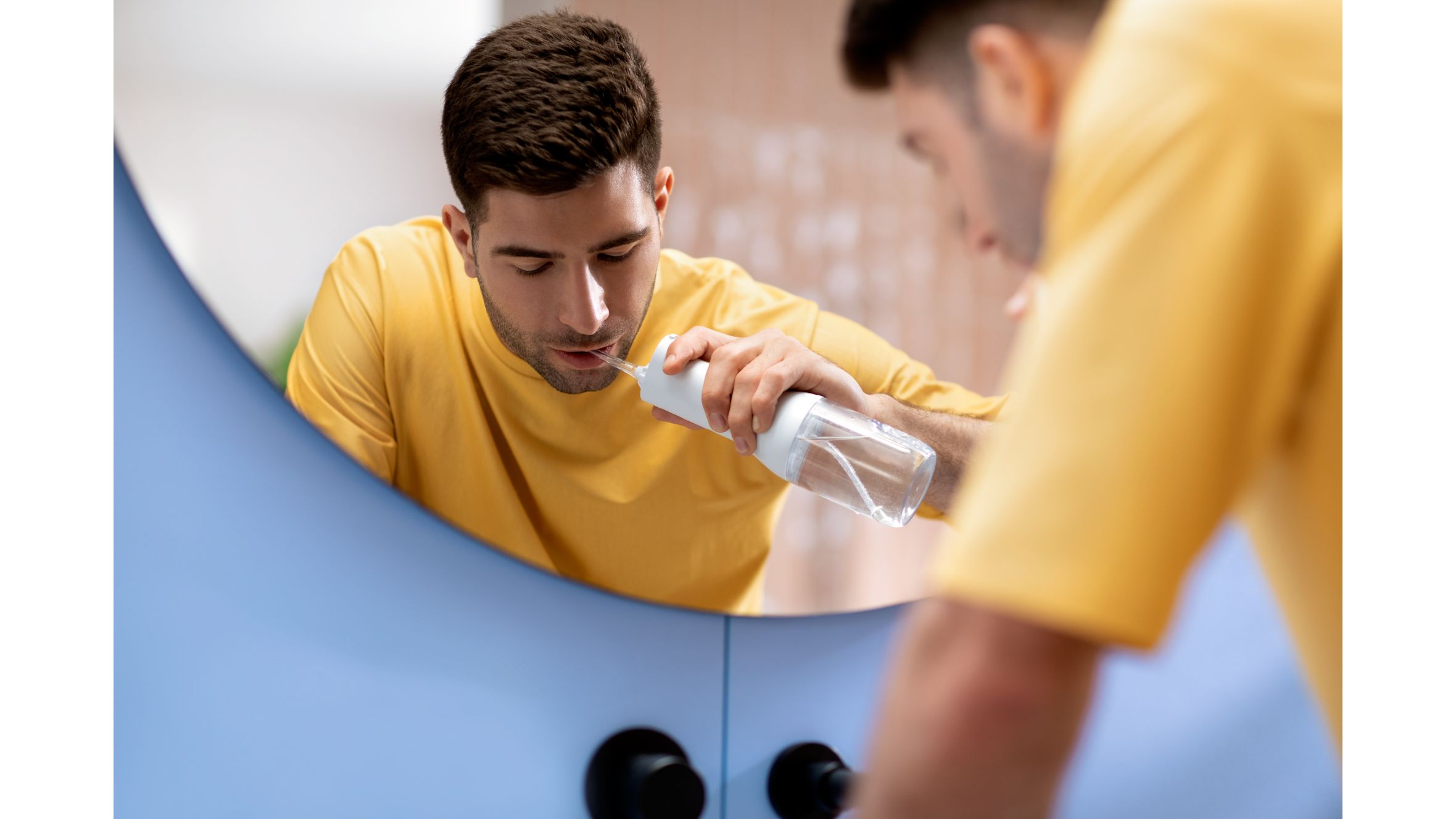 A young man in a yellow shirt bends over the bathroom sink while brushing his teeth with a Philips electric toothbrush.