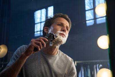 Hombre utilizando una afeitadora rotativa con espuma de afeitar frente al espejo. T&eacute;cnica perfecta para un afeitado apurado en mojado.