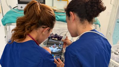 Two healthcare professionals review ultrasound images on a tablet beside an infant incubator.