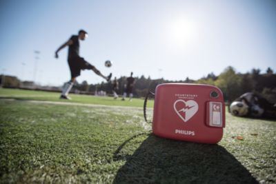 Philips-AED placerad på fotbollsplan med spelare i bakgrunden.