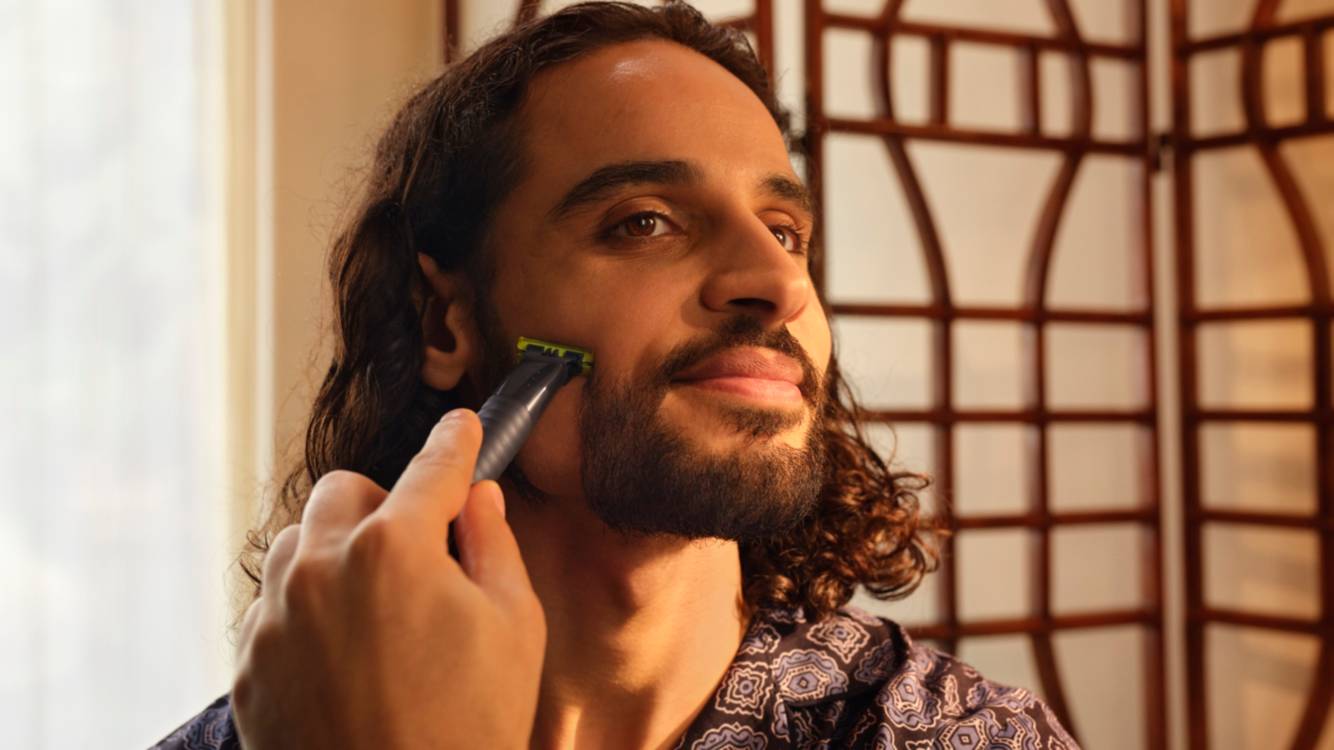 Ein Mann mit lockigem Haar lächelt, als er sich mit dem Philips OneBlade die Wange rasiert.