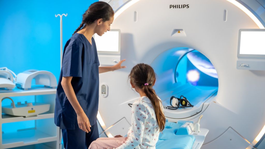 Pediatric Coaching: Girl on MRI bed, looking in the bore to Ollie content with nurse – In-bore Connect