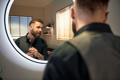 Un hombre con ducktail barba se mira en un espejo circular muy iluminado.