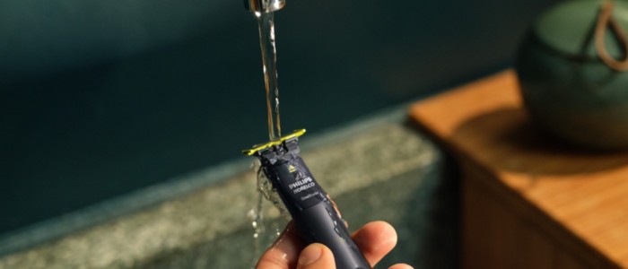 A Philips OneBlade shaver is held under a running tap, demonstrating its waterproofing.