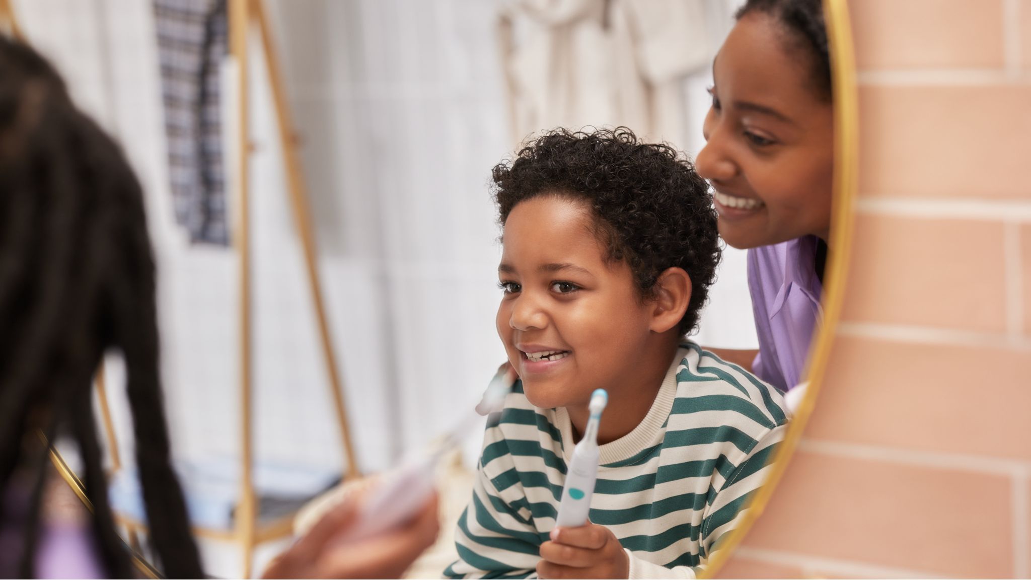 A young mother helps her child to brush their teeth correctly while looking in the mirror.