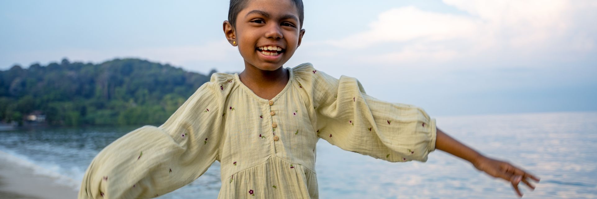 young child with arms open wide standing on sand with the ocean behind