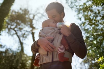 Elternteil füttert ein Baby mit der Flasche, während es das Kind nach draußen trägt