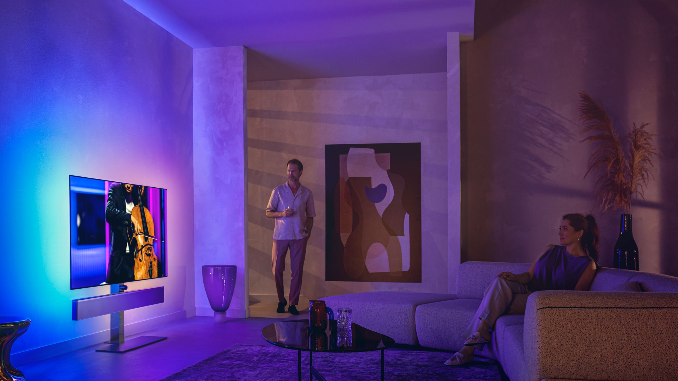 Man and woman in a stylish living room watching a Philips Ambilight TV displaying a cello performance