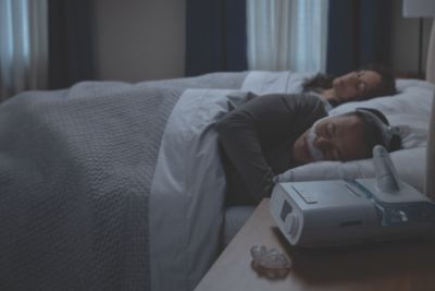DreamStation on Nightstand, Couple, Man wearing DreamWear Nasal sleeping in bed