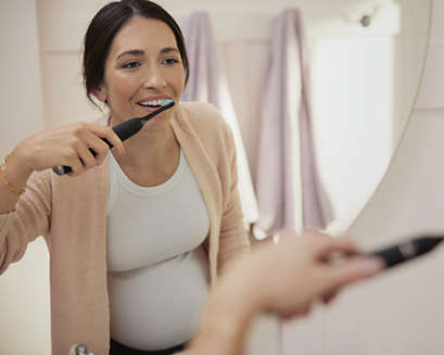 A pregnant women brushing her teeth and looking in a mirror.