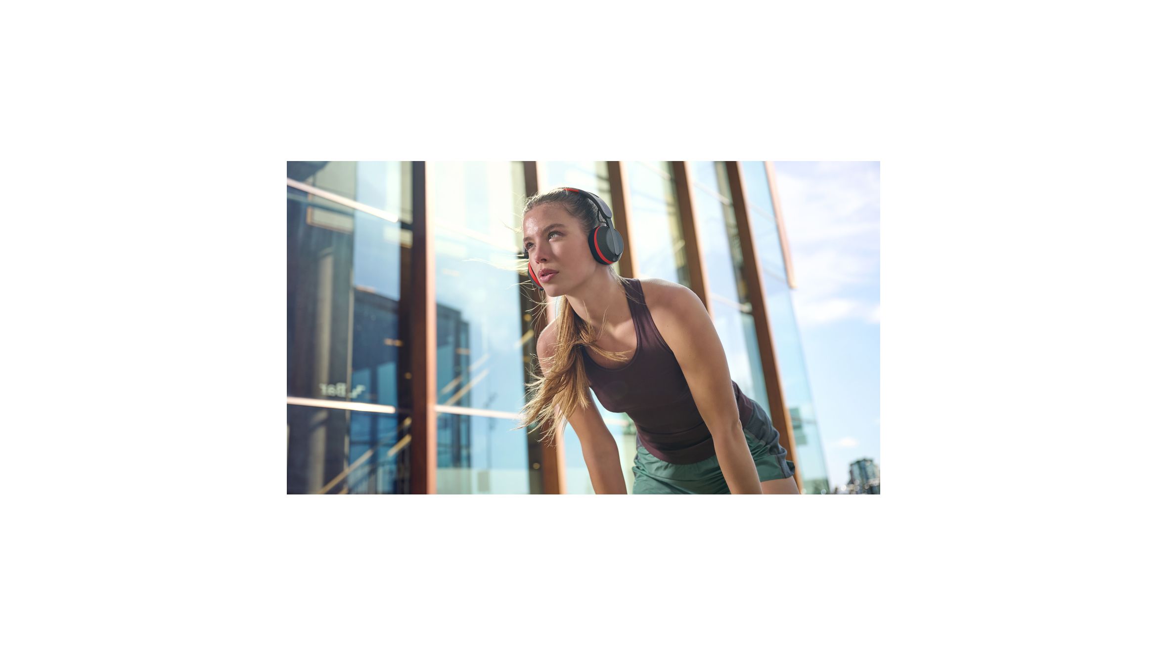 Une femme portant un casque audio Philips A6219 fait une pause pendant une s&eacute;ance d'entra&icirc;nement en ext&eacute;rieur, devant un b&acirc;timent moderne en verre