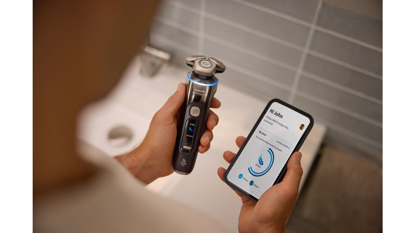 A man looking at the GroomTribe app on his phone while holding the Philips Shaver i9000 Prestige Ultra in the other hand.