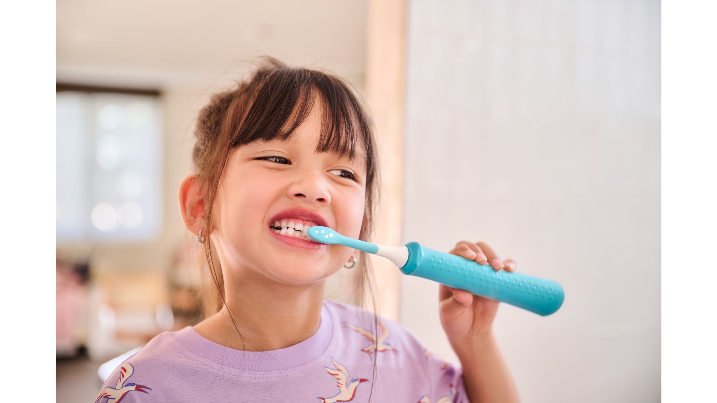 A young child wearing pink is shown brushing her teeth with a blue Philips kids electric toothbrush and kids toothbrush head.