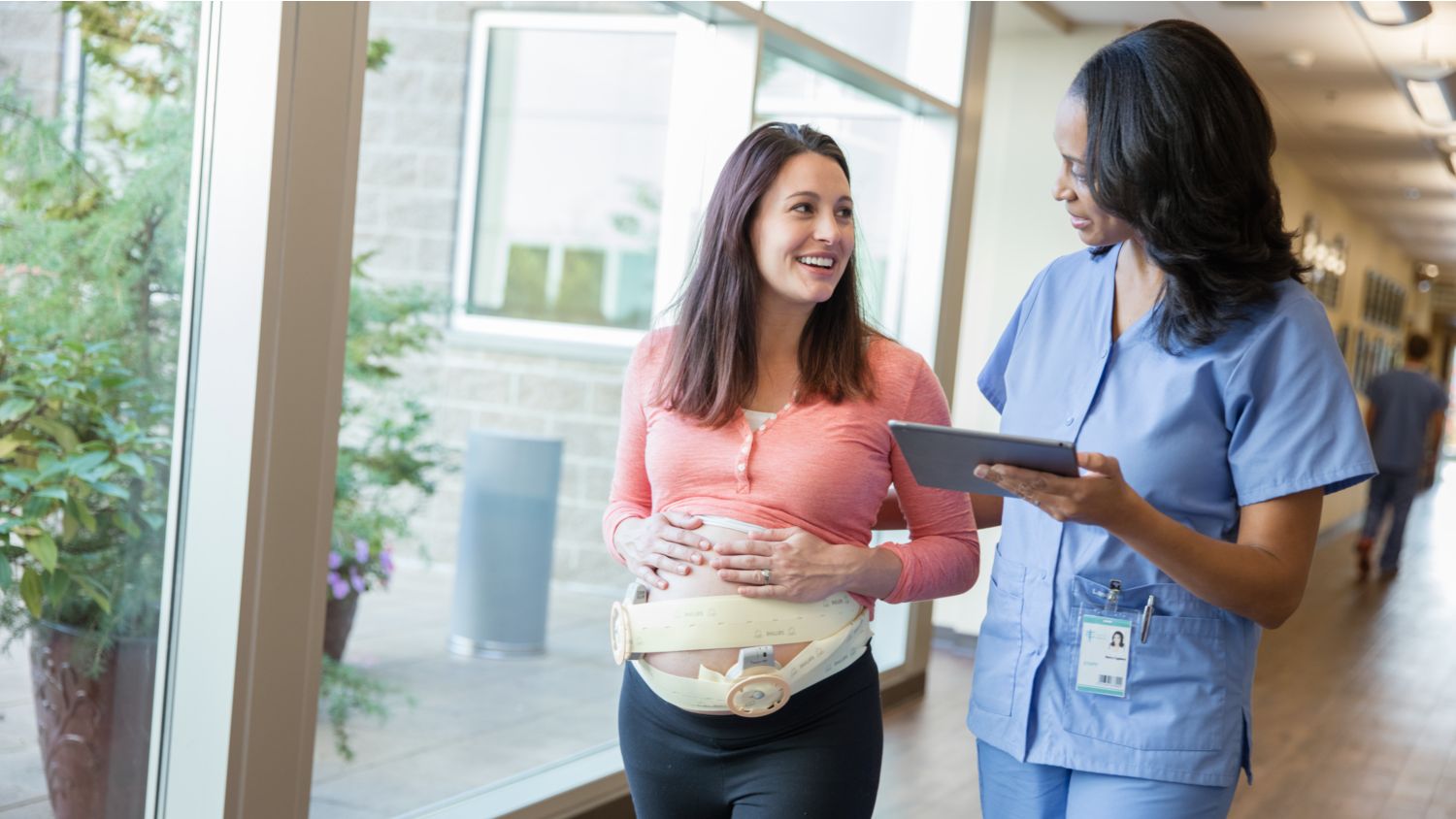 Woman wearing fetal monitor walking down hospital hallway with provider