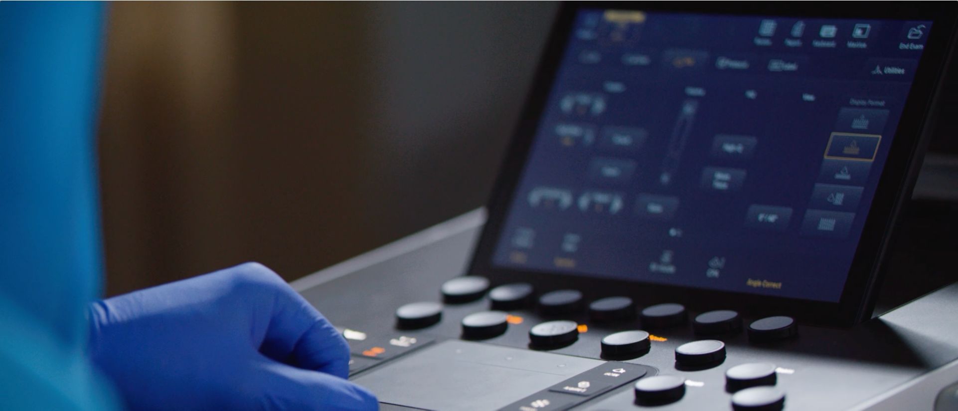 Mano con guante azul ajusta los controles de un equipo de ultrasonido; la pantalla del equipo aparece al fondo