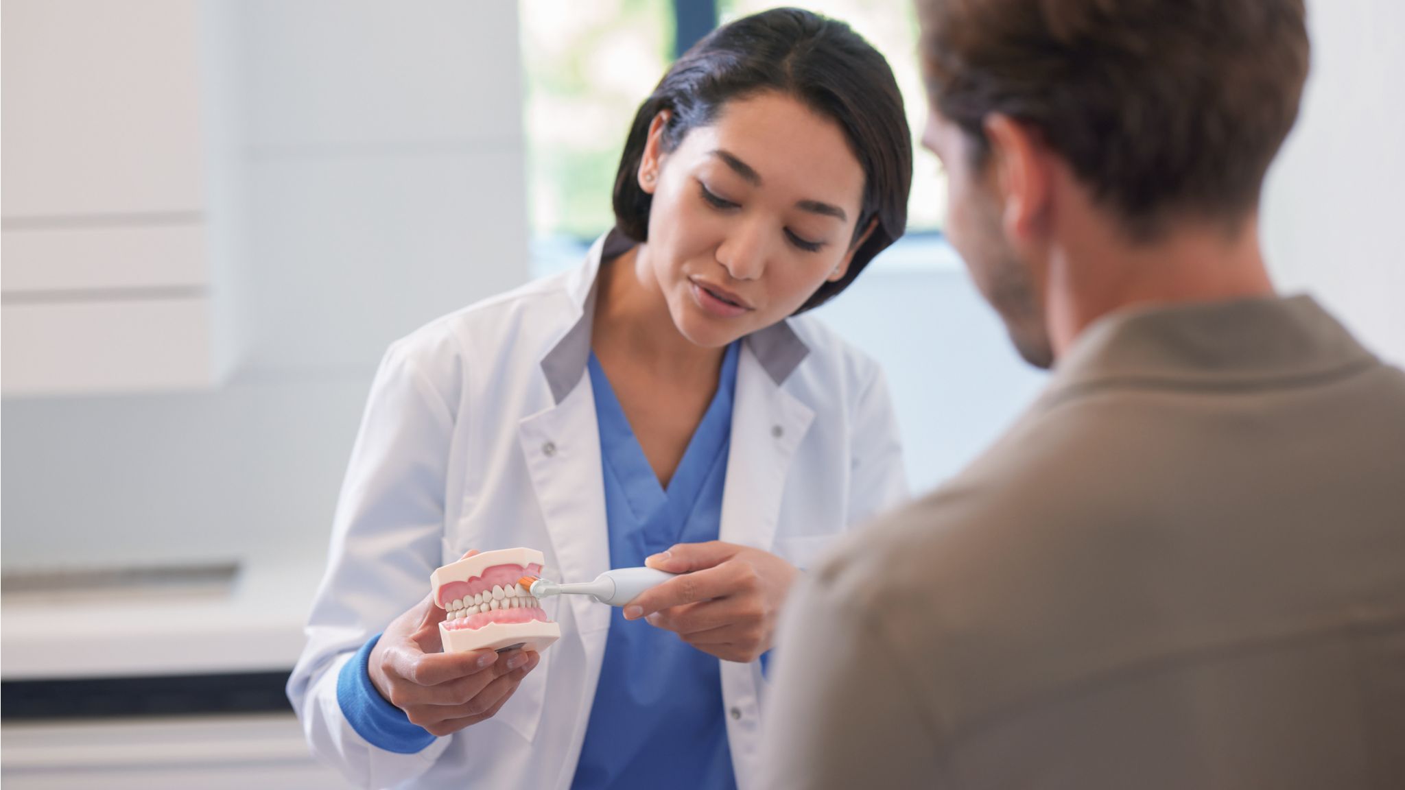 A young dental professional demonstrates professional dental cleaning to her patient using a model of the mouth and a Philips electric toothbrush.