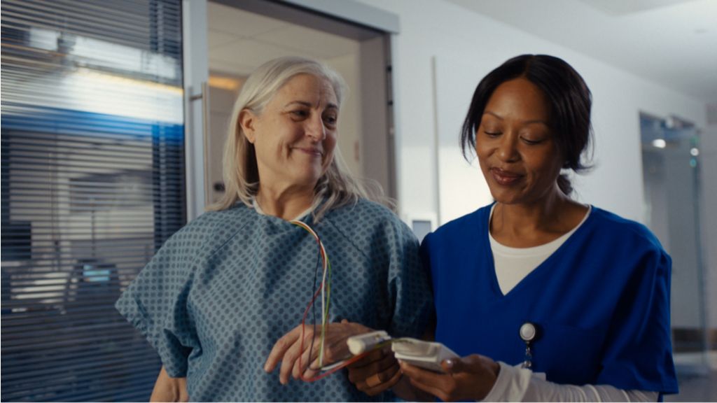 The patient is continuing her recovery as she walks in a hospital hallway with a clinician while being monitored by the Philips Telemetry Monitor 5500.