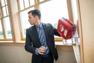 A man in an office setting takes a Philips HeartStart FRx AED from a wall mounted cabinet. The scene highlights emergency preparedness and access to defibrillation equipment.