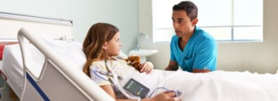 A healthcare professional in blue scrubs speaks with a young patient lying in a hospital bed, who is holding a stuffed toy and connected to a monitoring device.