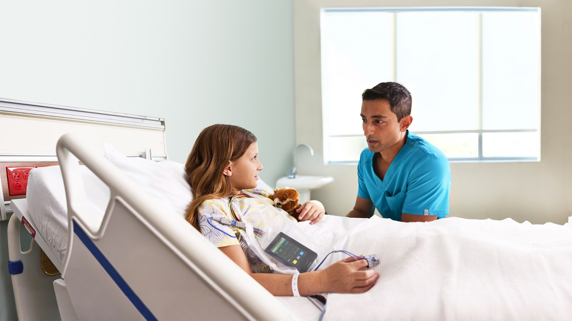 A healthcare worker speaks with a child patient in a hospital bed, with patient monitoring equipment visible