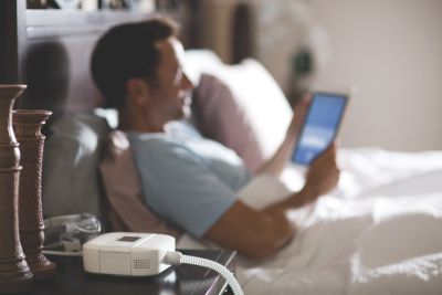 Person i säng som använder surfplatta med CPAP-enhet på nattduksbordet