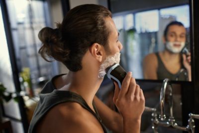 En mann med man bun-frisyre barberer det tykke skjegget sitt foran et baderomsspeil.