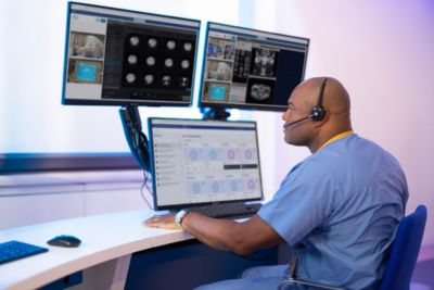 Remote MRI technologist reviewing brain scans and vital data on a triple‐monitor Philips workstation with headset