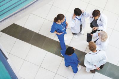 Widok z góry na niewielką grupę pracowników ochrony zdrowia (w strojach medycznych i białych fartuchach) stojących na szpitalnym korytarzu i prowadzących dyskusję. Jeden z klinicystów trzyma tablet/urządzenie mobilne
