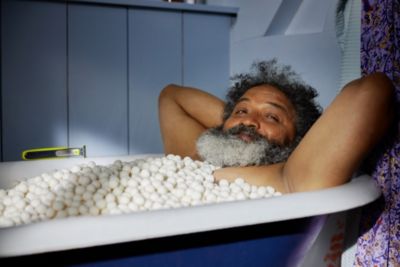 Un hombre con barba poblada se relaja en una ba&ntilde;era llena de guijarros blancos. Cerca hay una Philips OneBlade.