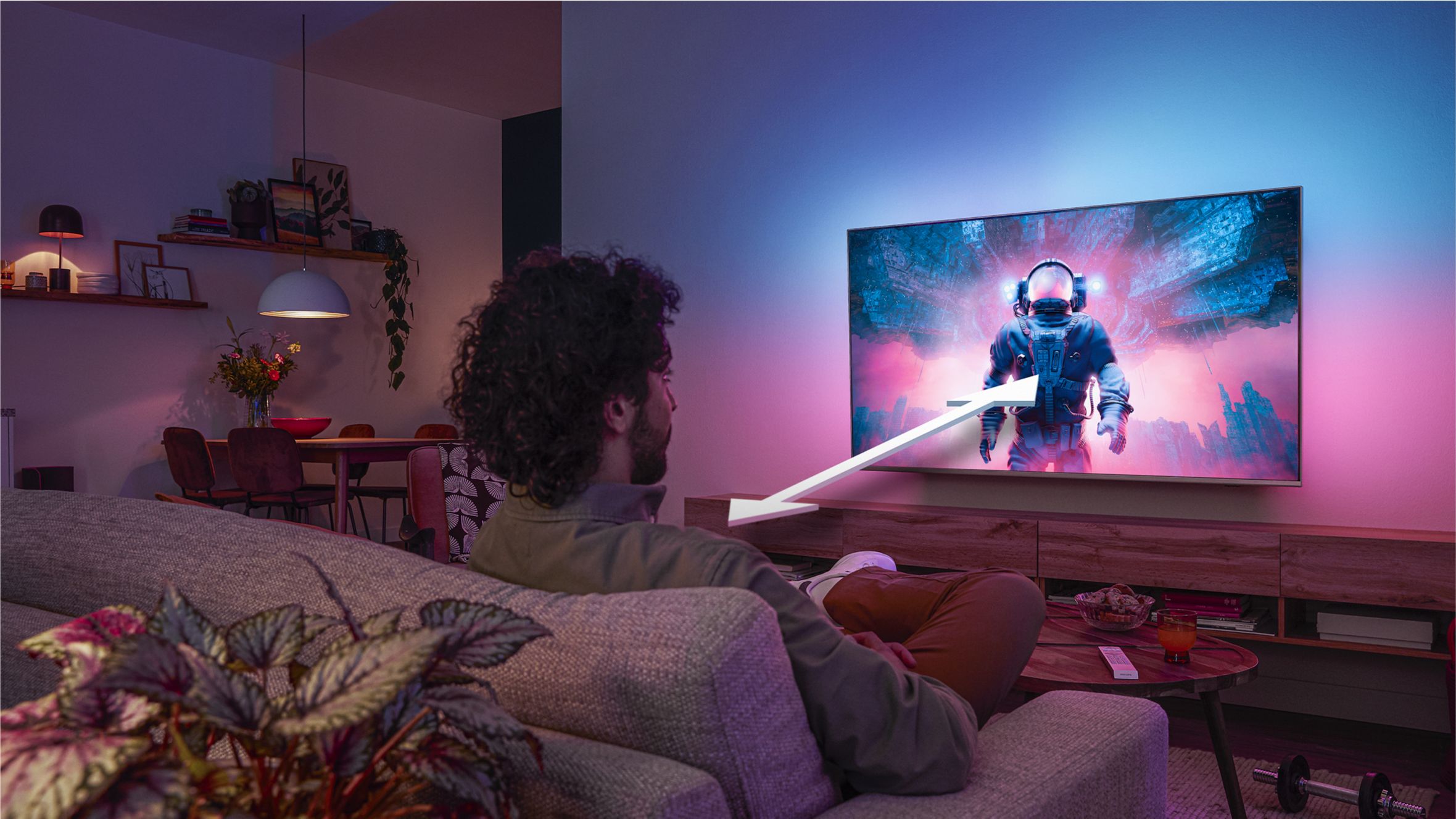Man sitting on a sofa watching a Philips Ambilight TV displaying a colorful astronaut scene, with an arrow indicating optimal viewing distance