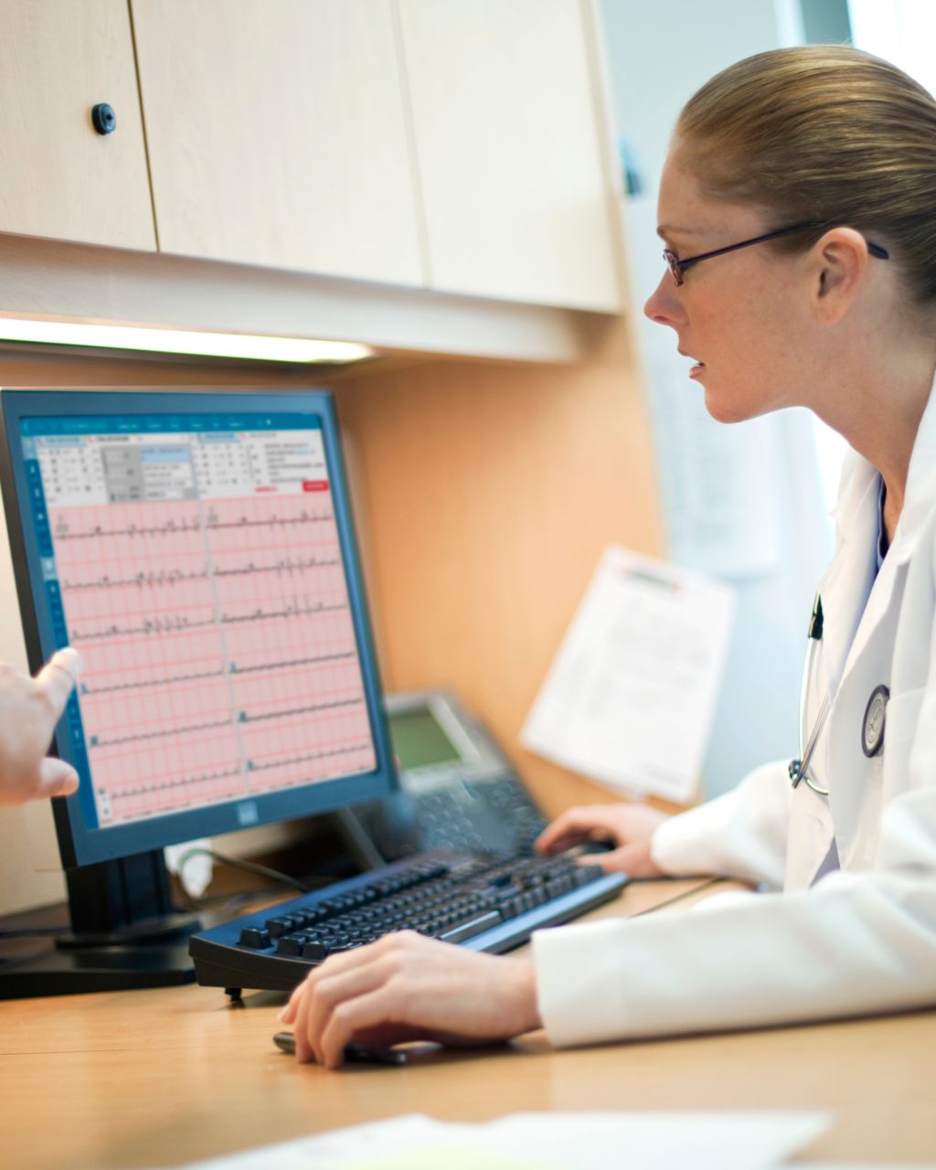 Two clinicians in white lab coats review ECG data on a desktop computer.