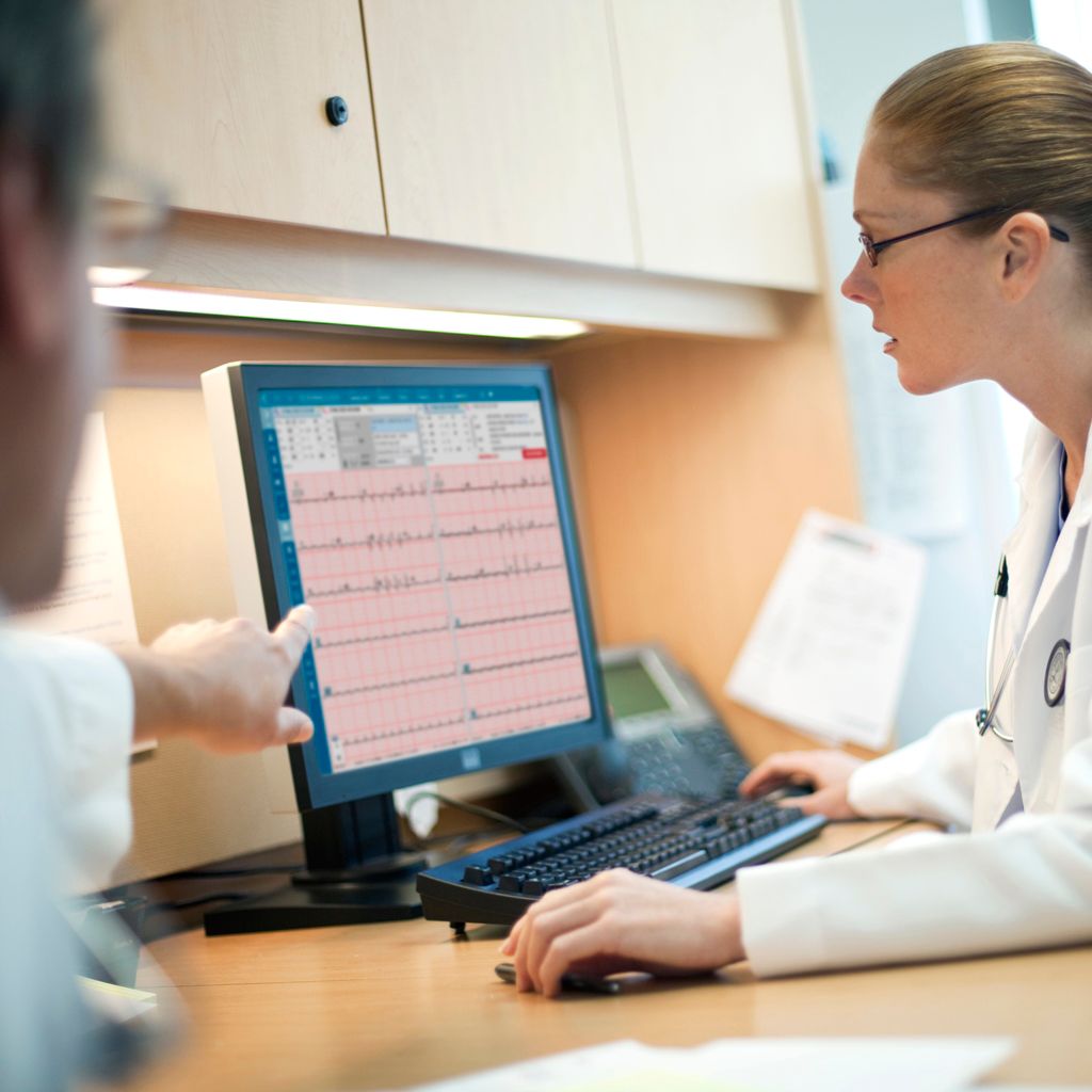 Two clinicians in white lab coats review ECG data on a desktop computer. 