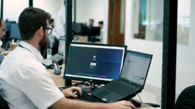 Profissional de TI da saúde trabalhando no sistema Tasy utilizando telas duplas e notebook em ambiente de escritório
