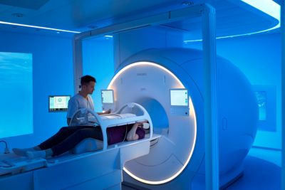 A radiology professional is preparing a patient lying on a table for an MRI scan using a Philips MR system in a room illuminated with ambient blue lighting. The MRI machine features a circular design with integrated display screens on both sides.