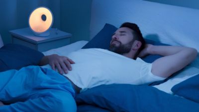 Mężczyzna śpiący w łóżku z lampką Wake Up Light świecącą na stoliku nocnym