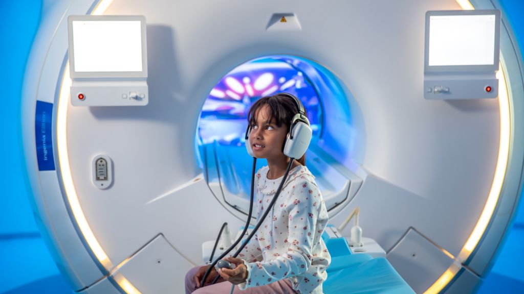 Girl sitting on MRI bed with headphones and controls.