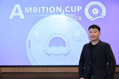 อุเทน ยารัตน์ ยืนอยู่ด้านหน้าป้ายจัดแสดงเครื่อง MRI ในงาน Ambition Cup 2024 ณ งานนวัตกรรมเทคโนโลยีด้านการดูแลสุขภาพ