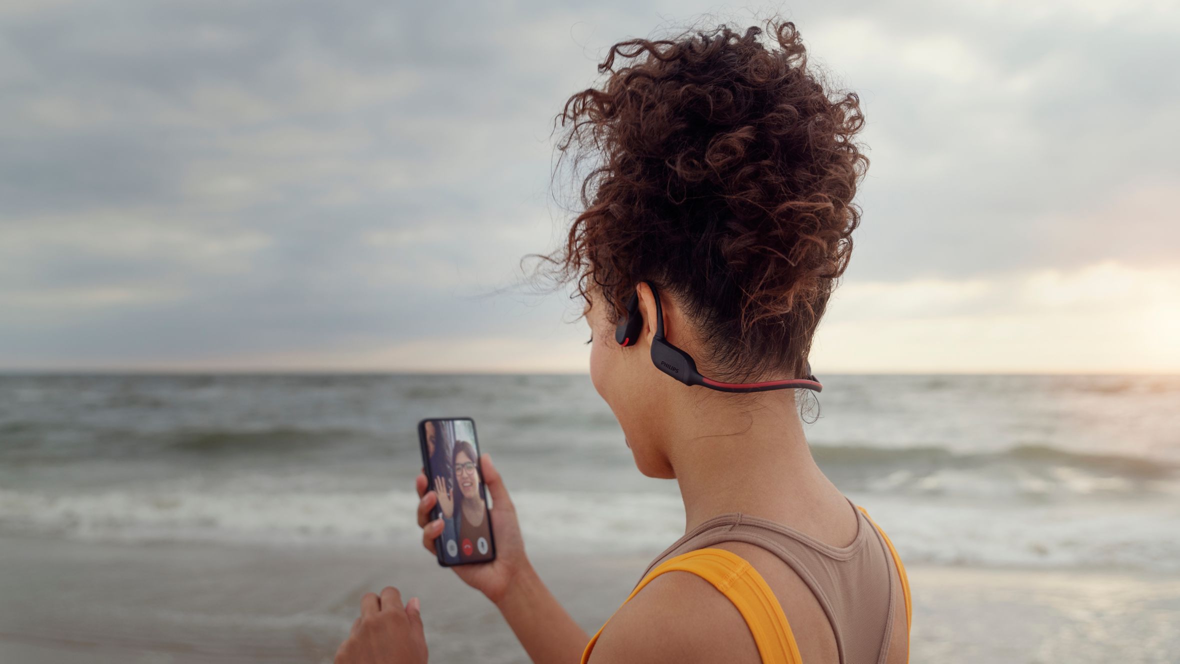 Sportol&oacute; telefon&aacute;l a strandon, mik&ouml;zben csontvezet&eacute;ses fejhallgat&oacute;t haszn&aacute;l