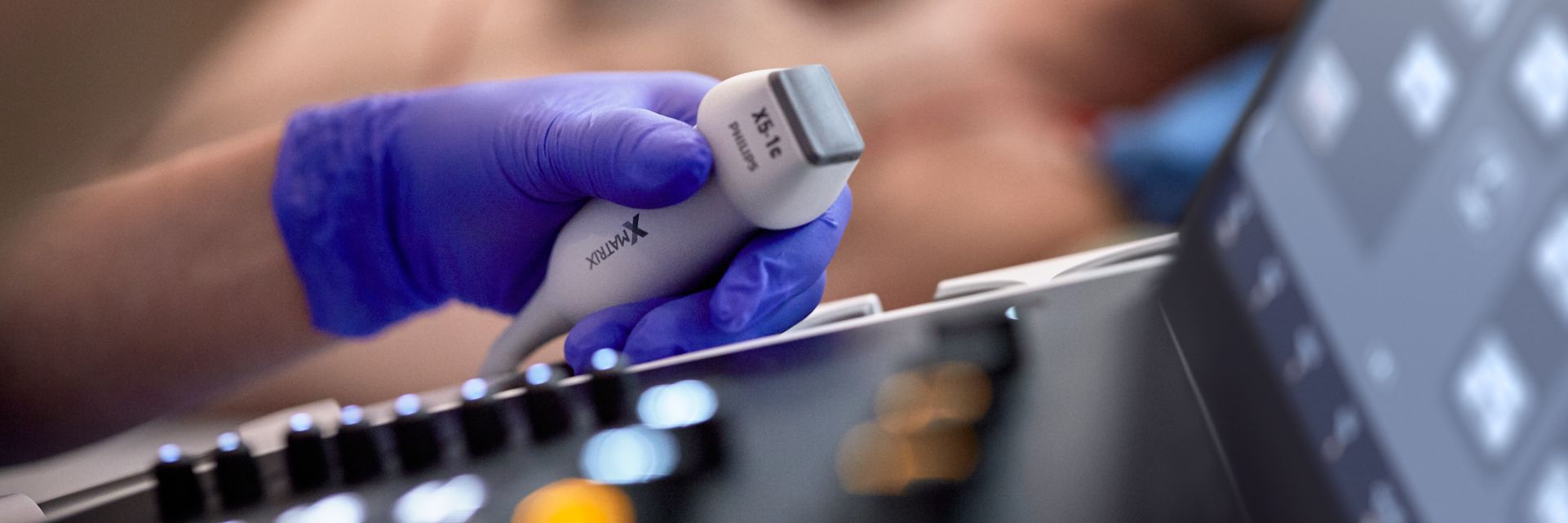 Primer plano de una mano con guante morado sosteniendo un transductor de ultrasonido sobre el panel de control de un ecógrafo, durante la realización de un examen médico