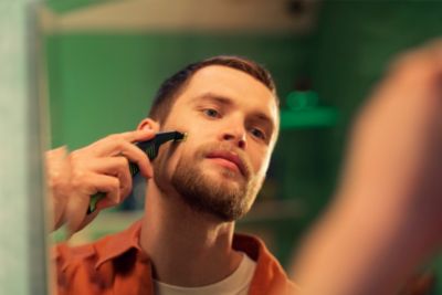 A man wearing an orange shirt shaves his clean beard with the Philips OneBlade in a bathroom mirror.