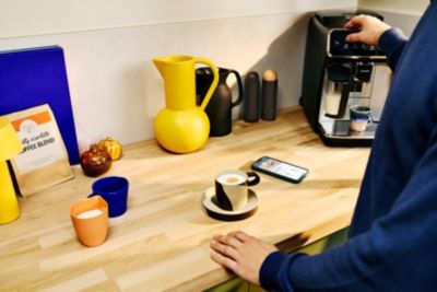 Person using a coffee machine via touchscreen; a cup, milk and a smartphone with app interface are on the kitchen counter