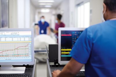 A medical professional monitoring patient data on two screens in hospital hallway