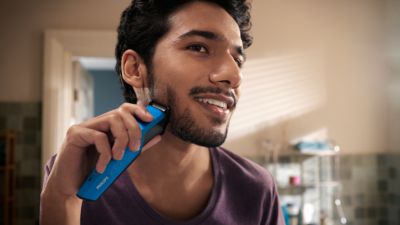 Un joven se mira de reojo en el espejo de un cuarto de ba&ntilde;o y se recorta la barba de la mejilla con una recortadora de barba Philips.