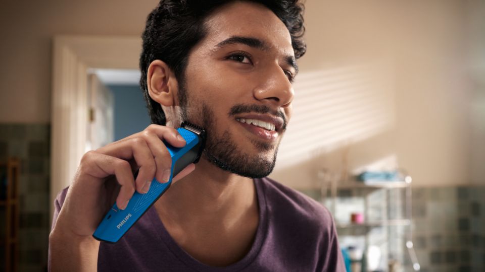 En ung man ler när han rakar sitt tjocka skägg med en elektrisk rakapparat från Philips.