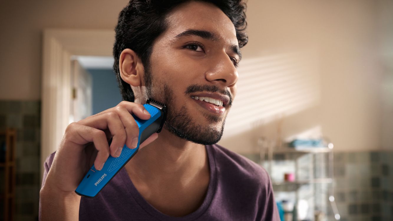 Ein junger Mann lächelt, als er seinen dicken Bart mit einem elektrischen Rasierer von Philips rasiert.