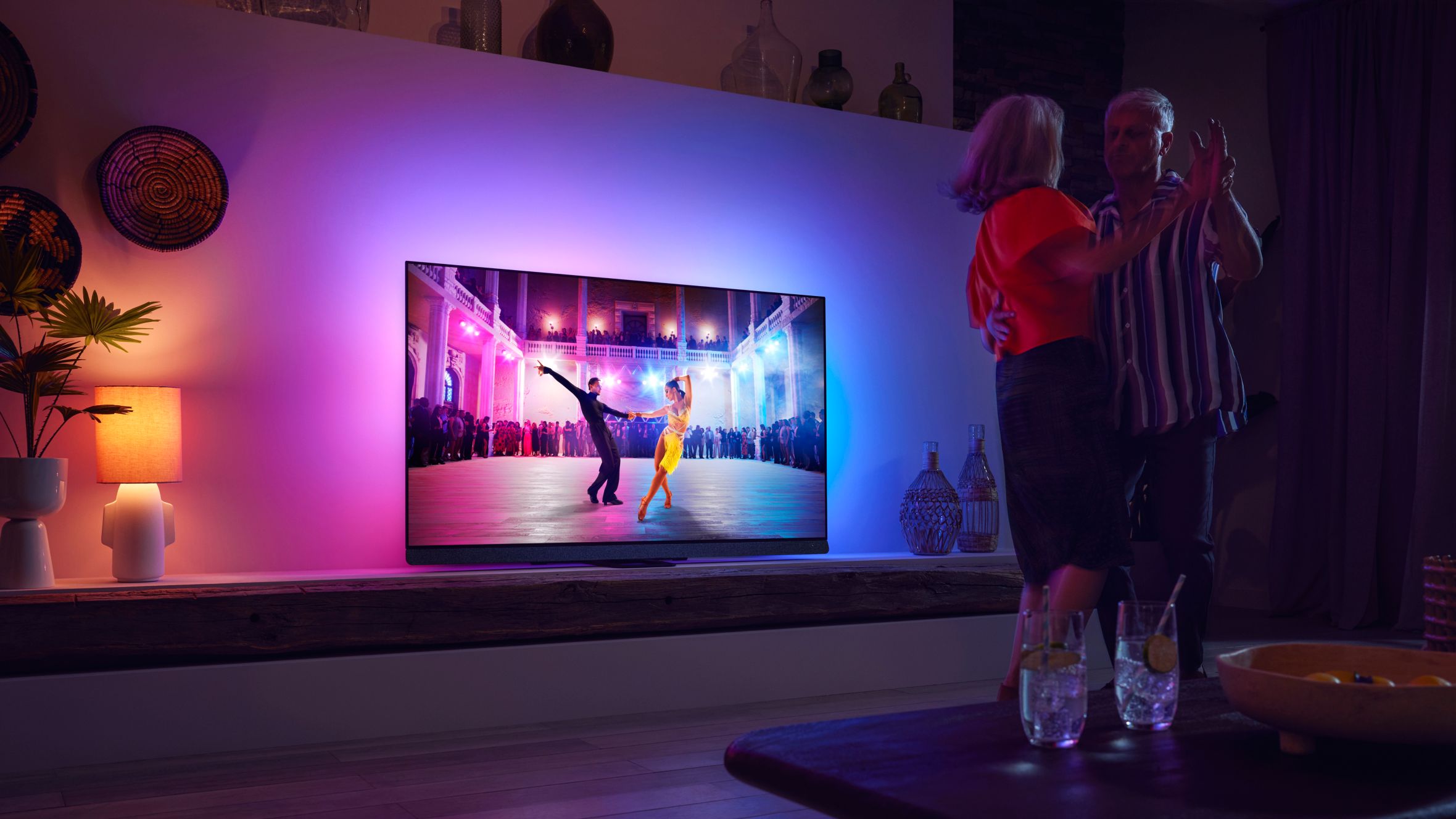 Couple dancing in a living room while watching a Philips Ambilight TV with colorful ambient lighting.