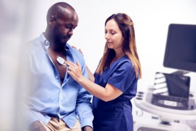 Nurse in blue scrubs applying wearable ECG sensor patch to male patient’s chest in exam room